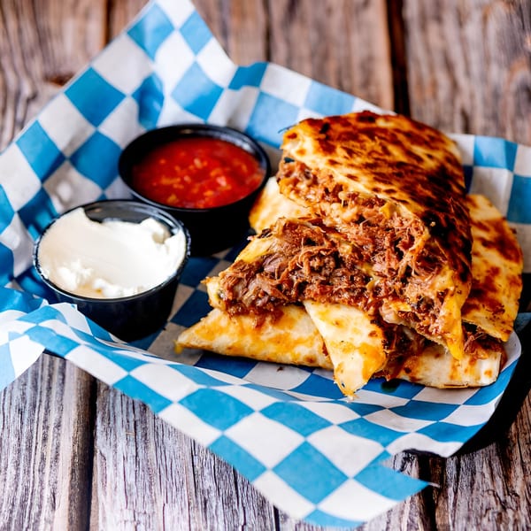Shredded beef quesadillas with salsa and sour cream in a blue and white checkered basket.