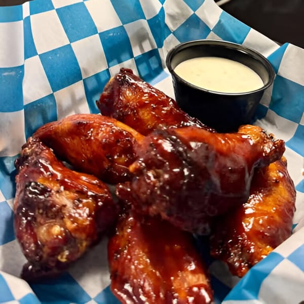 Glazed chicken wings with a side of dipping sauce in a blue and white checkered basket.