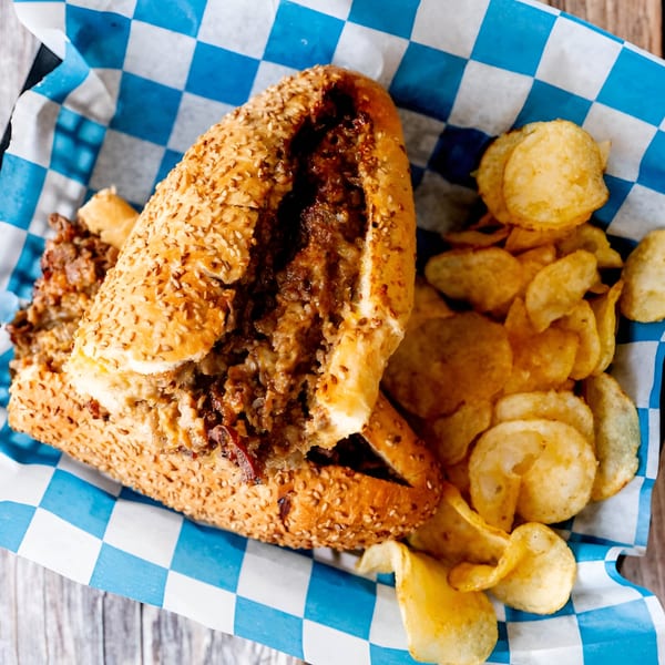 A Mac Daddies cheesesteak sandwich with potato chips in a blue and white checkered basket.